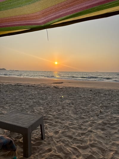 The image depicts a beach scene at sunset or sunrise in Bhagatwadi, India. The horizon divides a clear, warm orange and yellow sky, featuring a bright, low-angled sun slightly left of center, casting a luminous path across the water. The calm ocean exhibits gentle waves breaking on the shore, with several distant vessels silhouetted against the bright sky, including smaller boats to the left and a larger ship to the far right. The sandy beach in the foreground shows ripples and footprints, with some dark debris near the waterline. A dark gray rectangular plastic table with a textured surface rests on the sand in the lower left, partially obscuring a blue, green, and brown plaid fabric item. The scene is viewed from beneath a colorful, horizontally striped fabric awning or canopy (red, orange, green, yellow), from which a thin dark string hangs vertically. No individuals are explicitly visible, but the presence of the table and fabric suggests human activity or leisure.