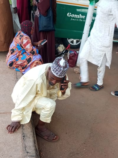 The image shows a street scene in Gombe, Nigeria, with a bus in the background that reads "Gombe Jewel In The Sa". In the foreground, a man in a yellow robe and patterned cap is kneeling and talking on a mobile phone. He is wearing brown sandals. To his left, a woman in a colorful patterned dress and headscarf is also kneeling and looking at her phone. Other people in traditional clothing are visible in the background. The ground is paved with a rough texture. The overall scene depicts a busy public space.