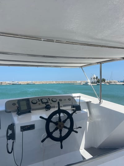 The image depicts the helm area of a boat, likely a yacht, with a clear view of the port area of M'Diq, Morocco. The boat's steering wheel is prominently featured, along with various control panels and gauges. A canvas awning structure provides shade. Outside the boat, the view includes the port's waterfront with buildings and infrastructure visible along the shore, and calm, turquoise water fills the harbor. No people are visible in the frame.