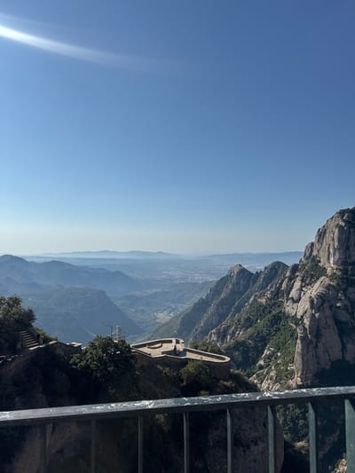 The image shows a panoramic view from a high vantage point, likely a mountain range near Collbató, Spain. In the foreground, a metal railing is visible, suggesting a viewing platform or balcony. Beyond the railing, a stone structure with a curved wall and steps leading up to it is built into the mountainside, surrounded by lush green trees. Further in the distance, rugged, rocky mountains dominate the landscape, interspersed with dense vegetation. The mountains recede into the background, with layers of hazy blue peaks stretching towards the horizon. A town or city is faintly visible in the valley below, and the sky is a clear, bright blue with a subtle lens flare in the upper left corner. The overall scene evokes a sense of natural grandeur and a popular tourist destination.