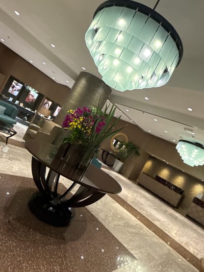 The image captures an interior scene, likely a hotel lobby or a public lounge area, viewed from an upward and tilted angle. The primary focus is a large, modern chandelier hanging from the ceiling, characterized by numerous translucent, light-colored (possibly frosted glass or plastic) rectangular panels arranged in a spiraling, tiered design. These panels emit a bright, cool-toned light from internal bulbs. Another similar chandelier is partially visible further in the background.
Below the main chandelier, a round table with a dark, sculpted base and a reflective glass top stands prominently. On the table, a vibrant floral arrangement features purple and yellow flowers mixed with long, green leaves, adding a splash of color to the setting.
The background reveals parts of the room's elegant decor. There are what appear to be multiple framed screens or artworks on a wall, showing images of people, though the details are not clearly discernible. Some light blue or teal upholstered seating can be glimpsed near these screens. A circular mirror with a golden frame is also visible on a wall further back, reflecting parts of the room. The ceiling is white with numerous recessed lights, and the floor is a light-toned, speckled material, possibly granite or marble, with contrasting darker striped patterns. The walls feature various textures and patterns, some reflective.
No people or identifiable activities are visible in the static shot. The lighting suggests an indoor setting, and no text is discernible within the image.