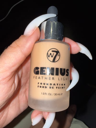In this close-up, indoor, still-life photograph, a person's hand, adorned with long, white acrylic nails, holds a bottle of W7 Genius Feather Light Foundation. The bottle is made of frosted glass and has a dropper applicator. The brand logo, "W7" inside a circle, is visible, along with the text "GENIUS" in large black lettering, followed by "FEATHER LIGHT," "FOUNDATION," and "FOND DE TEINT." Below this, the volume is listed as "1.0 Fl. Oz. / 30 mLe." The background is dark, suggesting low lighting conditions. There is no visible indication of the city of Odesa, Ukraine, or any outdoor urban environment.