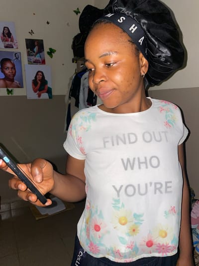 A young Black woman wearing a black satin bonnet and a white t-shirt with a floral print and the text "FIND OUT WHO YOU'RE" is holding a black smartphone in her left hand. She is looking down at the phone and smiling. The background includes a wall with several framed pictures and some clothing hanging on a rack. The floor is tiled. The image appears to be taken indoors, likely in a residential setting in Durumi, Nigeria.