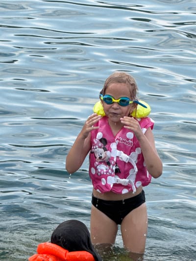 In this image, a young girl is in the water, wearing a pink and white polka dot life vest adorned with a Minnie Mouse print. She has on blue swimming goggles. Her black swimsuit bottoms are visible. Another person, whose head and shoulders are visible on the right side of the frame, is wearing an orange life vest. The water has a rippled texture, suggesting movement and light reflection. The scene appears to be outdoors, likely during the daytime, with the lighting and clear water indicating pleasant weather. The overall mood seems relaxed and playful, consistent with water activities. There is text visible on the life vest that reads "MINNIE". The location is identified as Partenit, Ukraine.