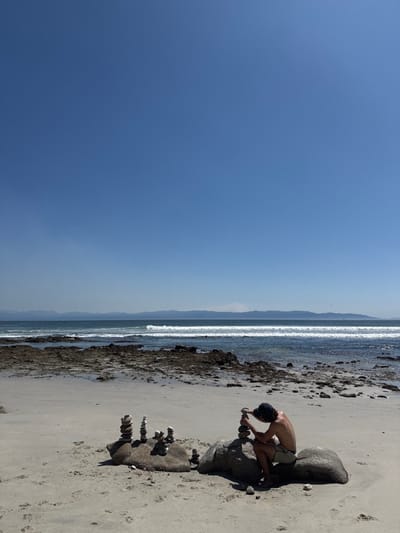 The image depicts a daytime scene on a sandy beach in Punta del Burro, Mexico. A single individual, appearing male, is seated on a large, smooth grey rock in the foreground, facing towards the left. He is shirtless, wearing light-colored shorts and a dark baseball cap, and is actively engaged in stacking small grey stones to form a cairn. To his left, another large, dark grey rock features several completed cairns of varying heights. The sandy beach extends towards the water, where a dark, rocky intertidal zone is visible. Beyond this, the ocean features light blue-green water with breaking white waves. A clear horizon separates the sea from a distant landmass or mountains under a bright, cloudless blue sky, which dominates the upper half of the frame. The overall environment suggests sunny weather.