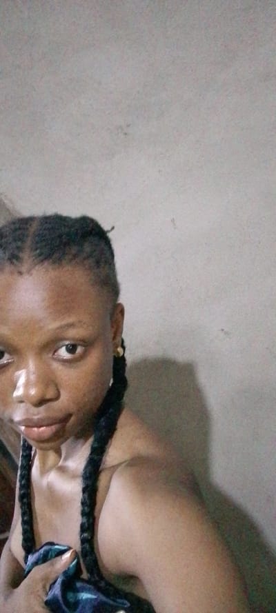 The image displays a close-up portrait of a young adult female with dark skin, situated indoors. She is positioned slightly off-center to the left, facing the camera directly with a neutral expression and a faint sheen on her lips. Her dark hair is styled in cornrows across her scalp, transitioning into long braids that extend over her shoulders; one prominent braid is visible over her left shoulder and another over her right. A small, circular, light-colored stud earring is present in her visible right earlobe. Her upper torso, including her shoulders and upper chest, is visible, suggesting she is wearing a dark garment or fabric, which her left hand is seen holding in the lower-left corner of the frame. The fabric appears dark with possible teal or green accents. The background is a plain, light grey wall, exhibiting subtle surface imperfections and a distinct shadow cast on the right side, indicating an off-camera light source. No specific landmarks are visible to confirm the location as Aba, Nigeria.