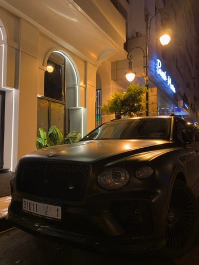 A dark-colored Bentley fronts a building with illuminated signage above. Streetlights cast a warm glow on the building's cream-colored facade, which features arched windows and architectural details. Plants decorate the exterior, adding a touch of greenery. The car's grill and headlights are prominently displayed, with a license plate visible on the front. The image captures the car at night, highlighting its sleek design against the backdrop of the building's architecture.