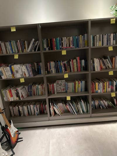 The image displays a brightly lit interior scene featuring three interconnected, floor-to-ceiling modular bookshelves, densely populated with books. The setting is identified as the Chongqing High-tech Zone, China.

The bookshelves are constructed from a gray, matte material, spanning the majority of the frame horizontally and extending upwards against a plain, light-colored wall. Each of the three vertical units contains four horizontal shelves, creating twelve distinct sections filled with books of varying sizes, colors, and thicknesses. The books are predominantly in Chinese script, with many standing upright, some leaning, and a few stacked horizontally.

Several yellow square sticky notes are attached to the front edges of the shelves, serving as labels.
*   On the top shelf of the leftmost unit, a sticky note reads "停止思考" (Stop Thinking).
*   On the third shelf from the top of the leftmost unit, a sticky note displays "阅读" (Reading).
*   On the bottom shelf of the leftmost unit, a sticky note reads "历史" (History).
*   On the top shelf of the rightmost unit, a sticky note shows "快乐" (Happiness).
*   On the second shelf from the top of the rightmost unit, a sticky note states "爱与" (Love and...).
*   On the third shelf from the top of the rightmost unit, a sticky note reads "艺术" (Art).
One book on the third shelf of the leftmost unit is open to reveal its cover, displaying the title "蔬菜教室" (Vegetable Classroom).

In the lower-left foreground, an electric guitar with a classic sunburst finish (transitioning from dark red to orange to yellow) and a white pickguard rests on a black metal stand. Only the upper body and neck of the guitar are fully visible. A second, partially visible white electric guitar is positioned further to the left, mostly out of frame.

The floor is a light gray, smooth surface, likely concrete or large tiles, with visible seams running horizontally and vertically. The wall above the bookshelves is uniformly light-colored, with a strong horizontal band of light visible near the top, indicating an overhead light source. A small cluster of green artificial foliage from a plant is visible in the top right corner, above the rightmost bookshelf unit.