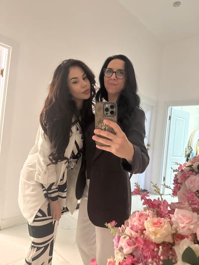 Two women pose for a selfie indoors, likely in a residence in Groningen, Netherlands. The woman on top, wearing black framed glasses, has long black hair and a dark blazer. She holds a smartphone with a patterned case. Below her, a younger woman with long brown hair wears a white and black patterned outfit. Behind them are white walls, a door with a white frame, and an arrangement of pink flowers.