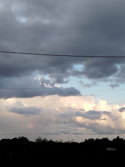 The image depicts a cloudy sky with dark storm clouds gathering overhead. Below the dense clouds, lighter, more textured clouds are illuminated by what appears to be sunlight breaking through. These lower clouds have a warm, pinkish-yellow hue, suggesting either sunset or sunrise. Several horizontal power lines stretch across the frame, dividing the sky into distinct sections. In the bottom portion of the image, a silhouetted treeline forms a dark, undulating horizon, obscuring any details of the landscape below. There is a faint, circular rainbow visible in the sky to the left of the center. The overall impression is one of a dramatic and somewhat ominous weather event approaching or passing. The time of day is likely late afternoon or early evening due to the lighting on the clouds. The location cues are limited to the power lines and silhouetted trees. No text is visible in the image.