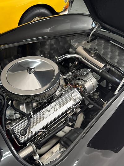 This image captures a detailed view of a car's engine bay, likely from a classic or custom performance vehicle, with a strong focus on the engine components. The primary subject is a V8 engine, featuring highly polished, finned chrome valve covers with "W/T" prominently displayed, suggesting a Chevrolet engine (possibly a small-block "World Tour" or a similar designation). A large, circular chrome air filter housing dominates the foreground, with its mesh filter element visible. To the left of the valve covers, a silver alternator with visible wiring is mounted.

The engine bay is meticulously presented, with various hoses, wires, and a chrome radiator overflow/expansion tank visible. Parts of the exhaust headers, wrapped in a white insulating material, can be seen in the background, contrasting with the dark engine block and other components. The inner fender and firewall panels are made of a textured, possibly brushed aluminum or diamond plate material.

The surrounding car body, particularly the fender and cowl, is painted in a glossy, dark grey or black finish, reflecting light. In the lower left quadrant of the image, a section of a bright yellow vehicle is visible, including a tire with "HANKOOK" faintly readable on its sidewall. This could be a spare tire mounted externally or part of an adjacent vehicle. A clear plastic tube, possibly for fluid overflow, is visible near the top left.

The scene appears to be indoors, possibly in a garage, showroom, or workshop, given the clean appearance and controlled lighting. No activity is taking place, and the car is stationary. The location context provided is Cape Town, South Africa, though no visual cues in the image directly confirm this.