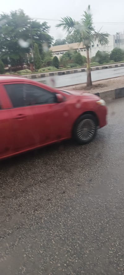 This image captures a red car driving on a wet asphalt road in Barakin Lerawi, Nigeria, under an overcast sky. The overall impression is of a rainy or recently rainy day, with visible water droplets on what appears to be the camera lens.
In the foreground, a bright red sedan is moving from left to right, exhibiting noticeable motion blur on its body and spinning blur on its wheels, indicating movement. The road surface is dark, wet asphalt, reflecting the dull light. A curb with black and white stripes separates the road from a green verge.
The background features abundant green trees and foliage, including a prominent palm tree on the right. Beyond the greenery, several buildings are visible; one appears to be a long, light-colored structure, possibly industrial or institutional, while another to its right is a taller, modern white building. The time of day appears to be daylight, despite the cloudy conditions. No people are visible, and there is no discernible text in the image.