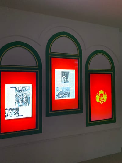The image shows three framed pictures mounted on a white wall in a room in Banguntapan, Indonesia, located at coordinates -7.8 latitude and 110.407 longitude. Each picture has a green frame with a gold trim and a red background. The leftmost picture displays a black and white photograph of a large group of people, possibly a crowd gathered for a public event. The middle picture displays a black and white photograph of several people standing together, possibly dignitaries or a group of individuals important to the local community. The rightmost picture features a gold emblem or crest on a red background, possibly representing a local organization or community. The room appears to be well-lit, with minimal shadows. There are no people present in the room, and no other objects or activities are visible. The wall surrounding the pictures is white and appears to be made of plaster or drywall.