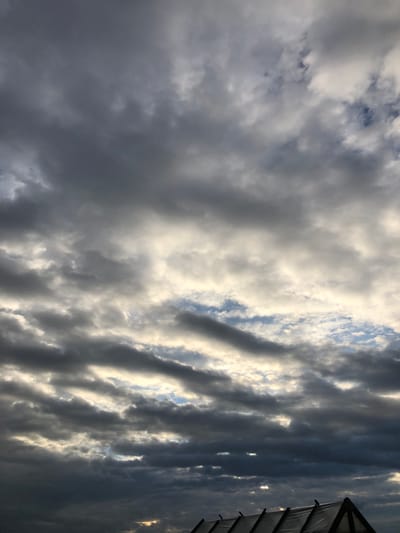 A low-angle shot captures a dramatic sky filled with an array of clouds. The clouds are a mix of dark, ominous storm clouds and lighter, broken clouds that allow glimpses of a bright, possibly sunlit sky behind them. This creates a sense of depth and dynamic lighting across the sky. In the lower portion of the image, a portion of a structure with a peaked roof is visible, covered with a translucent material. The scene appears to be an outdoor environment, potentially in the city of Uriupino, Russia, given the context. No people are visible in the frame, and no specific actions or interactions are depicted. The overall impression is one of an impending weather change.
