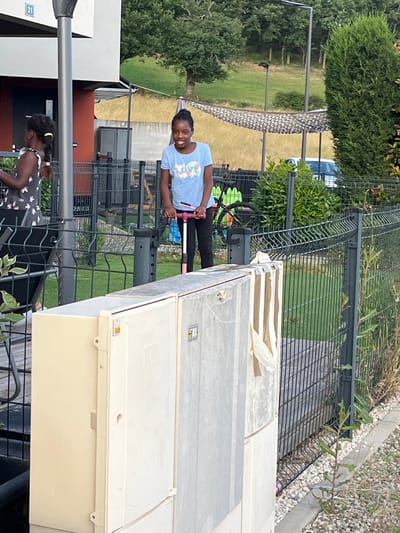 In Chambœuf, France, a young Black girl with braided hair stands on a pink and black scooter, smiling. She wears a light blue t-shirt with a colorful graphic and black pants. To her left, another young Black girl with braided hair in a white dress with a floral pattern is partially visible. They are in a fenced yard with green grass, trees, and a building with a red accent wall and a number "31" visible on a white facade. The background shows a wooded hillside and a white car. In the foreground, large cream and gray utility boxes are present. The overall scene depicts a residential outdoor setting on a sunny day.