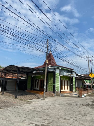 The image shows a mosque located at coordinates -7.498, 112.689 in Kecamatan Tanggulangin, Indonesia. The mosque has a green tiled exterior and a brown tiled roof with a small dome on top. The building is surrounded by a paved area and a small courtyard with a concrete ramp leading up to the entrance. Several power lines run across the sky above the mosque. There is a metal canopy covering an open garage area to the left of the mosque. In the background, there is another building and a power pole with several electrical wires. The sky is blue with white clouds. There are no people in the image.