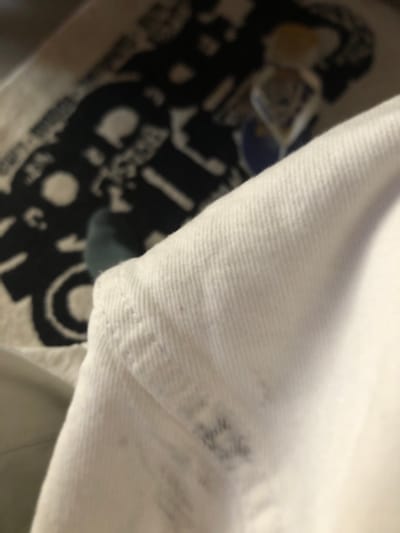 The image shows a close-up of white fabric, possibly denim, with some faint dark smudges on it. In the background, out of focus, is a dark pattern on a lighter surface, resembling a rug or tapestry. A person's foot, wearing a teal-colored sock, is partially visible in the lower left portion of the frame. The scene appears to be indoors, with no specific identifiable landmarks of L'Hospitalet de Llobregat, Spain, visible. The lighting is soft and indirect.