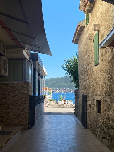 The image depicts a view through a narrow alleyway towards a body of water and a distant coastline. On the left, a building with a tiled facade and large glass windows is visible, partially covered by an awning. An air conditioning unit is mounted on the exterior wall. On the right, a building constructed of rough stone blocks features a green shuttered window and decorative stonework. A dark metal gate is present in the alleyway.
In the distance, a bright blue body of water stretches towards a hilly coastline dotted with buildings. The sky is clear and blue. On a paved area by the water, two individuals are casually dressed, suggesting a leisurely setting. The overall scene evokes a Mediterranean atmosphere, consistent with locations like Baošići, Montenegro, known for its coastal views and historic architecture.