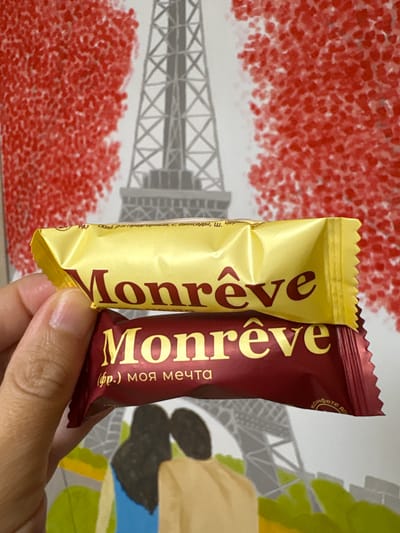 The image shows two candy wrappers held by a hand. Both wrappers have the brand name "Monrêve" printed in large gold letters. The top wrapper is yellow, and the bottom wrapper is dark red. The dark red wrapper also has Russian text below the brand name which translates to "(fr.) my dream". The background of the image features a stylized depiction of the Eiffel Tower with red, abstract shapes resembling foliage. The image is a close-up shot, focusing on the candy wrappers and the hand holding them. There is no visible indication of the location being in Vetluzhsky, Russia, or any other specific city.