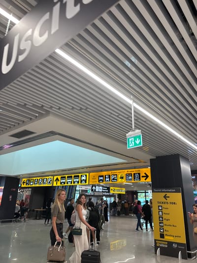 The image captures a scene inside a modern airport terminal, likely Fiumicino Leonardo da Vinci International Airport in Fiumicino, Italy, given the signage. The environment is spacious, with a high ceiling featuring ribbed metal panels and linear lighting. Large yellow directional signs are prominent, indicating various services like parking, train, taxi, bus, and hotel, with directional arrows. A green emergency exit sign with a person and down arrow symbol hangs from the ceiling.

In the foreground, two young women are walking, each with luggage. The woman on the left is wearing a khaki top and dark pants, carrying a light brown handbag. The woman on the right is wearing a white top and light-colored pants, with a small green purse. Several other people are visible in the midground and background, some walking, some standing, suggesting moderate airport activity. One of the yellow signs directs people to "Ai piani superiori - At upper levels". Another sign with "Tourist Information" lists "TICKETS FOR: Train, Hotel, Flight, Shuttle Bus, Bus, Meeting point" with corresponding icons and a directional arrow pointing left. The overall atmosphere is that of a busy but organized transportation hub.
