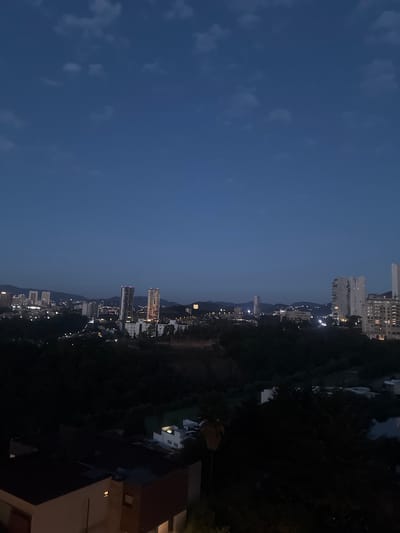 The image depicts a nighttime cityscape in Huixquilucan de Degollado, Mexico. Tall buildings are visible, illuminated by city lights, and contrasted against the dark sky and the foliage. The sky is a deep blue hue, suggesting either early evening or late night. The presence of buildings indicates a densely populated urban area. The image is vertically oriented.