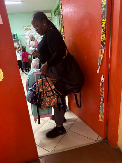 A woman in black stands beside a bright orange doorway adorned with a "Welcome" banner. She has braided hair and wears dark pants and shoes. Beside her are bags, including one with an orange fox pattern and a blue suitcase. In the background, a classroom setting shows students, desks, and wall decor, including a green board. The room is filled with activity and appears to be a vibrant learning environment.