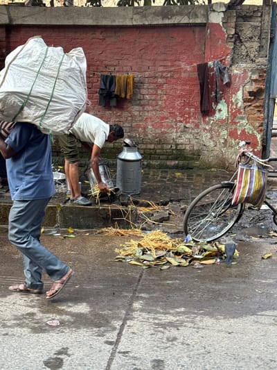 A man in a white shirt and khaki shorts is filling two metal milk cans with straw. He sits on a large white bag carried by another man in a blue shirt and gray pants walking along a street. A bicycle with a striped basket is leaning against a brick wall. Scattered debris and leaves are on the wet pavement.