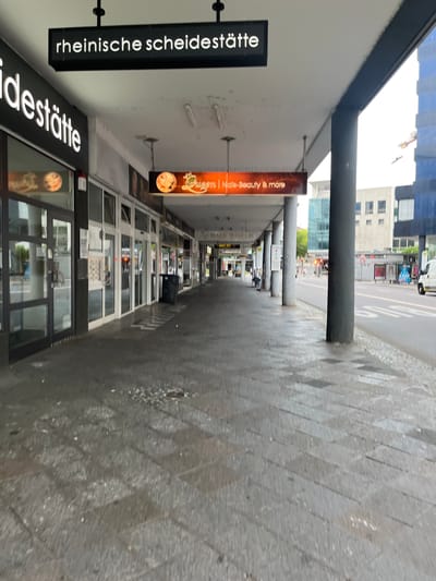 The image depicts a street scene in Saarbrücken, Germany, likely in a commercial area. A covered walkway runs along the left side of the image, with several storefronts visible behind glass doors and windows. One prominent sign above the walkway reads "rheinische scheidestätte" in white, illuminated lettering against a black background. Below this, another sign for "ideestätte" is also visible. Further down the walkway, a smaller, orange sign displays "Queen | Nails-Beauty & more" along with a graphic. The walkway floor is paved with large, dark gray tiles, which appear wet in places, suggesting recent rain or cleaning. A black trash bin is situated near the middle of the walkway.
The street itself is visible to the right, with a white van parked on the side and a bus stop shelter with advertising panels. Pedestrians are present on the sidewalk across the street, and traffic signals are visible. The background includes modern buildings, one of which is a tall blue glass structure. The overall impression is an urban environment, possibly during the day, with overcast or rainy weather conditions influencing the appearance of the pavement.