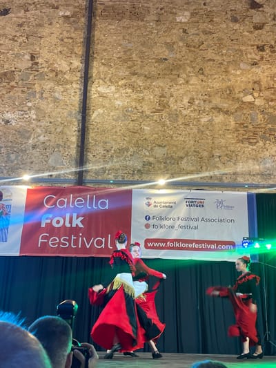 This media file captures a vibrant folk dance performance during the Calella Folk Festival in Calella, Spain. The scene is set on a stage with a backdrop displaying a large red banner that reads "Calella Folk Festival." The banner also includes logos and text related to the event's organizers, such as "Ajuntament de Calella," "FORTUNI VIATGES," and "Folklore Festival Association," along with a website address: "www.folklorefestival.com."
The primary subjects are two dancers dressed in traditional, brightly colored folk costumes. One dancer, in the foreground, wears a striking red dress with black accents and a white sash, her skirt dramatically flared. The other dancer, positioned higher in the frame, also sports a red and black ensemble. Both dancers appear to be in motion, their poses suggesting they are performing an energetic dance. Their expressions are focused, and the overall atmosphere conveyed is one of lively entertainment and cultural celebration.
The setting features a stone wall on the left side of the frame, suggesting an outdoor or historical venue. The stage itself is lit with colorful stage lights, including green and blue hues, which illuminate the dancers and the banner. In the bottom right corner, parts of the audience are visible, including hands holding cameras, indicating that the event is being photographed or filmed. The image is oriented vertically, likely captured by a person standing and filming or photographing the performance. The time of day is not explicitly stated but the artificial lighting suggests an evening or indoor event.