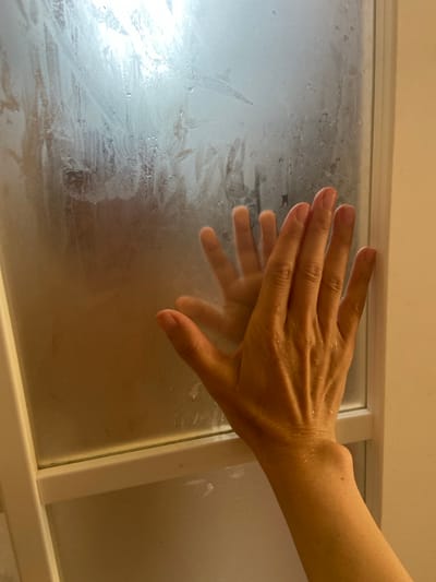 An arm and hand are pressed against a steamy glass shower door. The hand is in focus, with water droplets visible on the skin. The arm extends out of frame to the right. The glass is heavily fogged, obscuring the view of what is behind it, though a faint human silhouette can be discerned. The overall scene is dimly lit, with light sources appearing to be behind the shower door and from the side, illuminating the steam. The image is taken from a low angle, looking up towards the hand. There is no information in the image to suggest a specific location in the city of Tyumen, Russia.