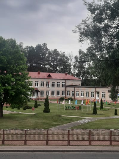 This is a medium-shot, vertically oriented photograph of a school building in Parechcha, Belarus. The building, made of light-colored bricks with a red-tiled roof, has multiple floors and numerous white-framed windows. In front of the school, a neatly mowed green lawn is adorned with small, colorful flags on poles. A paved pathway runs through the lawn, marked with white lines. To the right of the lawn is a wooden fence, and beyond that, a paved area that appears to be a road or sidewalk. Tall, dark green coniferous trees are planted strategically around the school grounds, and the surrounding area is filled with mature deciduous trees with lush green foliage. The sky is overcast and grey, suggesting a cloudy day. There is no visible activity or people in the photograph. The visible text on a green sign reads "УЖАКАР".