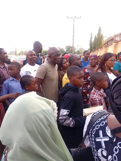 A diverse group of adults and children are assembled in an outdoor setting, possibly in Barden Yana, Nigeria. Most are looking in the same direction. A beige wall with barbed wire is visible on the right. A pole is visible in the background. A vehicle is visible in the distance. People are wearing a variety of clothing.