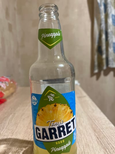 The image shows a partially empty glass bottle of "Tony's GARRET Hard Pineapple" drink. The bottle is clear glass with a green and blue label featuring a pineapple slice and text. The bottle is placed on a light-colored wooden surface, possibly a table. In the background, a beige-colored wall and a curtain with a blue and white checkered pattern are visible. The scene is indoors, and there are no people or animals present. The image does not contain any information that would allow for the determination of its location in the city of Domodedovo, Russia, or any other specific location.