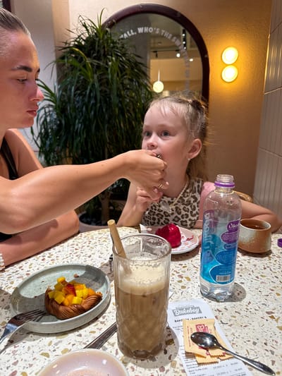 In a brightly lit cafe, possibly located in Nha Trang, Vietnam, a woman is feeding a piece of food to a young girl. The woman, seen from the side, has her arm extended towards the girl, who is looking at the food with her mouth open. The girl has blonde hair in a ponytail and is wearing a leopard-print top. On the table in front of them, there is a pastry topped with fruit, a glass of iced coffee with a bamboo straw, a bottle of water, a small cup of tea, and a receipt from "Bakeboy - Bakery &". The cafe's interior features a large potted plant and a wall with textured tiles. A round mirror with text is visible in the background, and two circular lights are mounted on the wall. The table surface is speckled with various colors.