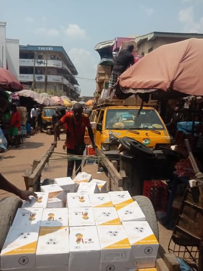 The image depicts a busy street market scene in Ogidi, Nigeria, under a bright sky. In the foreground, a wooden handcart is laden with approximately thirty white cardboard boxes, each bearing the brand "NWYCON MEGA" and illustrations of a child with a pencil, a power outlet, or an insect symbol. A person's left forearm and hand are visible on the far left, near the front wheel of the handcart. A man in a red-orange shirt and dark pants, with a dark bag slung across his chest, pulls the handcart by its wooden handles, facing forward.
Immediately behind the handcart and to its right is a yellow commercial van, heavily loaded with various items, including boxes and a large, dark, urn-shaped object on its roof rack. A man in a dark t-shirt and plaid shorts is positioned on top of the van, actively arranging or securing the cargo. A large, reddish-brown market umbrella partially obscures the view on the right side of the midground.
The background shows a bustling street with multiple market stalls, indicated by various open umbrellas. Several pedestrians are visible, along with a yellow three-wheeled auto-rickshaw (keke Napep). On the left, a multi-story building with "TECNO" branding and "SPARK 40" signage is visible. The ground appears to be unpaved, consistent with a market thoroughfare. The sky is light blue with scattered white clouds, suggesting daylight conditions.