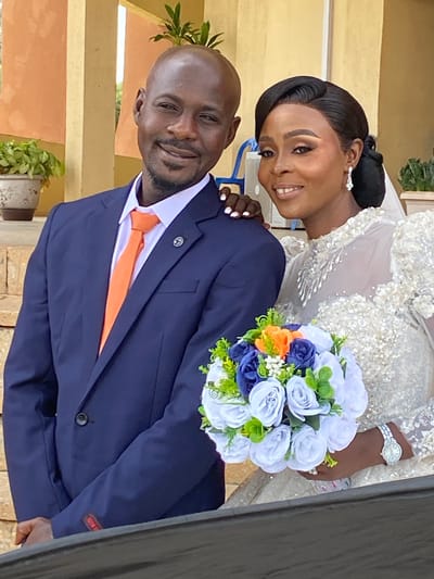 This image captures a wedding portrait of a Nigerian couple, likely taken in Kuru Karama, Nigeria. The bride, adorned in an exquisite white wedding gown with intricate beadwork and lace detailing, leans towards her groom. Her hair is styled in an elegant updo, complemented by pearl earrings. She wears a watch on her left wrist and a ring on her right hand. The groom, dressed in a sharp navy blue suit, white shirt, and an orange tie, smiles warmly at the camera. He has a boutonnière pinned to his lapel. The couple is holding a beautiful bouquet of flowers, featuring shades of white, blue, and orange roses with green foliage.
The background suggests an outdoor or semi-outdoor setting, with hints of yellowish walls and terracotta-colored planters containing greenery. The lighting indicates it's daytime. The overall mood is joyful and celebratory, as evidenced by their smiles and embrace. The image is oriented vertically, with the couple positioned centrally.