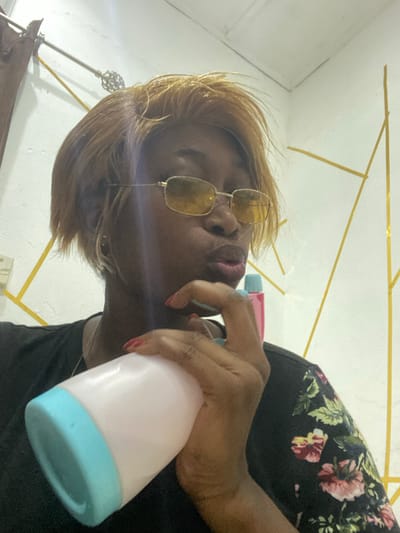 A tilted selfie features a person with blonde hair and gold sunglasses, making a duck-face expression. They are holding a pink and blue spray bottle. The individual is wearing a black floral shirt. A decorative gold-painted geometric design is visible on the white wall behind them. The image is brightly lit and angled upwards, giving a distorted perspective. A decorative lamp or stand is partially visible in the background.