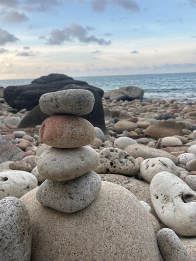 The image depicts a rocky beach in Baan Chaloklum, Thailand. The foreground showcases an arrangement of five smooth, oval-shaped stones, oriented vertically. These stones vary in color, including shades of gray and reddish-brown. They rest atop a bed of similar rocks and pebbles. In the background, the ocean is visible under a cloudy sky. A dark, possibly volcanic rock formation is positioned near the horizon, separating the stones in the foreground from the ocean. There are no people or identifiable subjects other than the rocks and the landscape. The interaction present is the arrangement of the stones. The environment is a natural beach setting at dusk or dawn.