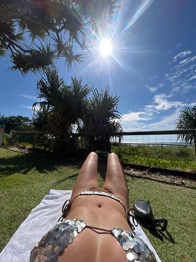 The image displays an outdoor scene captured on a bright, sunny day, with a vertical composition. A person, likely female, is shown lying on a white towel on green grass, extending from the lower-right foreground towards the middle-left of the frame. The person has tanned skin and is wearing a brown bikini embellished with numerous large, round, metallic silver-colored sequins or disks on the straps of both the top and bottom. A black shoulder bag or small purse rests on the grass near the person's upper body in the upper-right portion of the image.
The background features a vibrant blue sky with scattered white, wispy clouds. The sun is prominently visible in the middle-left, emitting a strong sunburst effect with rainbow lens flares, indicating a direct shot into the sun and clear weather. A metal fence runs vertically through the midground, separating the grassy area from dense, green, tropical-looking vegetation, including trees with spiky leaves that resemble Pandanus. Beyond this foliage, a narrow strip of sparkling ocean is visible, suggesting a coastal setting, consistent with Coolangatta, Australia. The foreground consists of well-maintained green grass, exhibiting clear shadows cast by the sun. Faint and unreadable informational signs are partially visible in the lower-left background. The overall impression is one of leisure and relaxation in a beautiful, sunny, coastal environment.