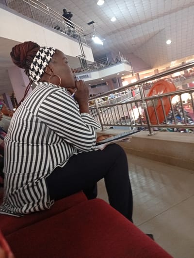 A woman in a black and white striped shirt and black pants is sitting on a red seat. She has brown braided hair with a black and white headband. She is wearing hoop earrings and has her hand to her chin as if in thought. She's holding a tablet or book. The scene appears to be indoors, perhaps in a large auditorium or church with a railing and other people in the background. A person can be seen on a balcony.