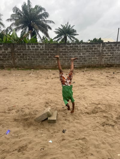 A young person is performing a handstand on a sandy surface in Badagry, Nigeria. The person is wearing a green shirt and patterned shorts. Two rectangular concrete blocks are situated to the left of the handstand. A grey brick wall forms a backdrop, with palm trees and vegetation visible behind it. The ground is covered in sand with scattered debris, including plastic items. The sky is overcast.