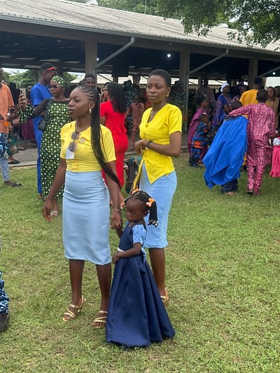 This image depicts an outdoor gathering in Itoga, Nigeria. In the foreground, two adult women and a young girl are the primary subjects. The woman on the left wears a yellow short-sleeved top and a light blue skirt, accessorized with sunglasses on her chest and gold strappy sandals. She holds the hand of the young girl standing to her right, who is dressed in a dark blue dress with puffed sleeves and bows in her braided hair. The woman on the right wears a yellow top and a light blue skirt, similar to the first woman's outfit, and also sports gold strappy sandals. She holds her left hand by her side and her right hand is not visible.
The background reveals a crowd of people dressed in various colorful attire, suggesting a festive occasion. Several individuals are visible under a large, open-sided structure with a corrugated roof, possibly a pavilion or community hall. The ground is covered in green grass, and trees and foliage are visible in the distance, indicating a rural or semi-rural setting. The overall atmosphere appears to be one of community and celebration.