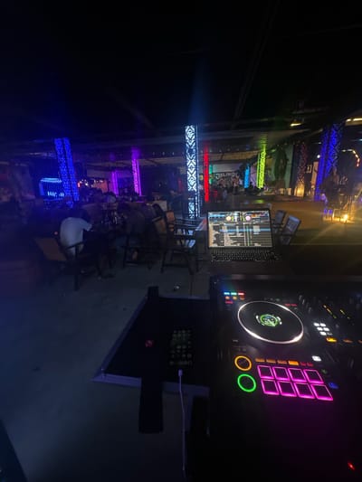 This image depicts an outdoor evening scene, likely in a commercial area in Howayi, Nigeria. In the foreground, a DJ setup is visible, featuring a laptop connected to a DJ controller with illuminated pads and a large jog wheel. The DJ controller is predominantly black with vibrant pink and green lights.
The background reveals a lively atmosphere with a crowd of people seated at tables, suggestive of a restaurant or bar. The area is illuminated by colorful, decorative lights, including tall, illuminated pillars displaying intricate patterns in blue, purple, red, and green. In the far background, a sign with text, possibly a business name or event title, is partially visible, reading "JALINGO".
The ground appears to be paved, and various seating arrangements, including wooden chairs and tables, are scattered throughout the space. The overall impression is one of a social gathering taking place in a well-lit, entertainment-oriented venue at night.