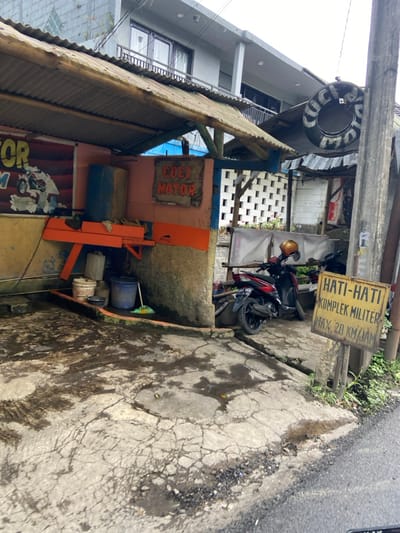 The scene shows a motorcycle parked outside a building in Lembang, Indonesia. The building has a sign that reads "Cuci Motor," indicating it is a motorcycle wash. The motorcycle is red and black, with a yellow helmet on the handlebars. There is a yellow sign with Indonesian text, likely a warning sign, next to the motorcycle. In the foreground, there is a muddy and cracked concrete area, likely a parking lot or driveway. There are also some buckets and other cleaning supplies visible. The image was captured at the coordinates -6.825 latitude and 107.61 longitude.