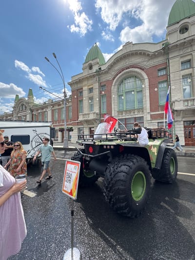 In a sunlit street scene in Novosibirsk, Russia, a large, camouflage-patterned all-terrain vehicle (ATV) with oversized tires is the central focus. The ATV, which appears to be a specialized expedition or off-road vehicle, is parked on a wet asphalt surface, indicating recent rain or street cleaning. Two Russian flags are prominently displayed on the vehicle, one a large tricolor and another smaller red flag, suggesting a patriotic or event-related display.
Several people are visible around the ATV and in the background. In the foreground on the left, a person in a light purple top holds a white cup. To their right, a woman in a floral dress and sunglasses stands near a white recreational vehicle (RV) adorned with a compass graphic labeled "ROAD TRIP". A man in a light blue polo shirt and denim shorts walks past them. Further in the background, a woman in a white shirt is seated on the ATV, and another person, possibly a man, is visible behind her. A few other individuals are also seen in the distance, including a person walking away from the ATV towards a building.
The backdrop consists of an ornate, historic building with red brick facade, arched windows with green frames, and decorative elements, characteristic of architectural styles found in cities like Novosibirsk. The building features green domes and what appears to be the year "1910" inscribed above a large archway, hinting at its age and historical significance. Streetlights and electrical wires crisscross the sky, and scattered white clouds are present in the bright blue sky. A sign with a QR code and Russian text is placed in front of the ATV, likely for promotional or informational purposes. The overall scene suggests an outdoor event or exhibition in a city center.