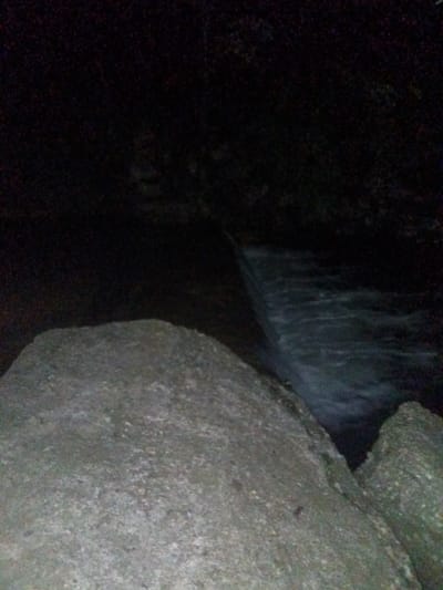 The image displays a dark, grainy night scene of a natural landscape, likely taken in Tahlequah, USA. The foreground is dominated by large, textured rocks or boulders, appearing in shades of grey and dark brown, occupying the lower half of the frame. These rocks show some rough texture and granular detail, hinting at a natural, rocky environment.

Behind the prominent rocks, a body of water, possibly a river or stream, flows. The water appears dark but shows signs of movement, with blurred ripples and slight reflections indicating its current, possibly forming a small waterfall or rapids as it descends slightly to the right side of the image.

The background is almost entirely enveloped in deep darkness, making it difficult to discern specific features. It appears to be an indistinct mass, possibly representing distant land, trees, or rock formations, all obscured by the lack of light.

The photograph was taken at night or very late dusk, as indicated by the extreme low light conditions and the overall blackness dominating the scene. There are no people, specific activities, or visible text in the image. The general atmosphere is one of quiet, dark natural beauty.