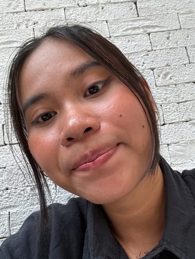 A close-up, high-angle selfie of a young woman with dark hair and brown eyes is shown. She is wearing a black collared shirt. Her face is tilted slightly, and she is looking towards the camera with a slight smile. Her skin appears to have some blush applied to her cheeks. She has two moles on her right cheek. The background is a textured white brick wall. The lighting suggests an outdoor setting, possibly during the day. The location is South Jakarta, Indonesia.