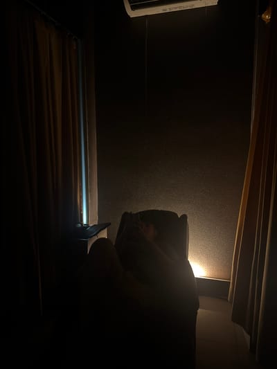 The image shows a dimly lit interior space. A person is seated in what appears to be a chair, though only the upper portion of their body is visible and largely obscured by shadow. The background is a dark wall, possibly textured. Below the wall is a horizontal surface, lit by a faint light source, and covered with what appear to be folded fabrics. The location is Gunung Putri, Indonesia.