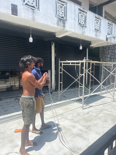Two men are working outdoors in the city of Weligama, Sri Lanka, in front of a building with a stone facade and black roll-up doors. The man in the foreground, shirtless and wearing grey shorts, holds a long metal pole. He is looking to the right, towards the second man who is wearing a blue shirt and tan shorts. The second man is also looking to the right, appearing to assist with the pole. They are standing on a concrete surface, with scaffolding visible to their right. A white pendant light hangs from an overhang above the roll-up doors. The area appears to be undergoing some form of construction or renovation.
