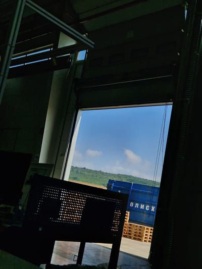 The image depicts the interior of a large industrial building, possibly a warehouse or a factory, with a large loading bay door open to the outside. The interior is dark and industrial, with exposed metal beams and structures. In the foreground, a dark, perforated object, possibly a piece of furniture or equipment, is silhouetted against the bright exterior. Through the open doorway, a bright blue sky with scattered clouds is visible, along with a green, tree-covered hillside in the distance. A blue shipping container with white Cyrillic lettering, "ОЛИСХ," is parked outside, along with several wooden pallets. The scene suggests a logistics or transportation hub. The location is specified as Bahovitsa, Bulgaria. There are no people visible in the image.